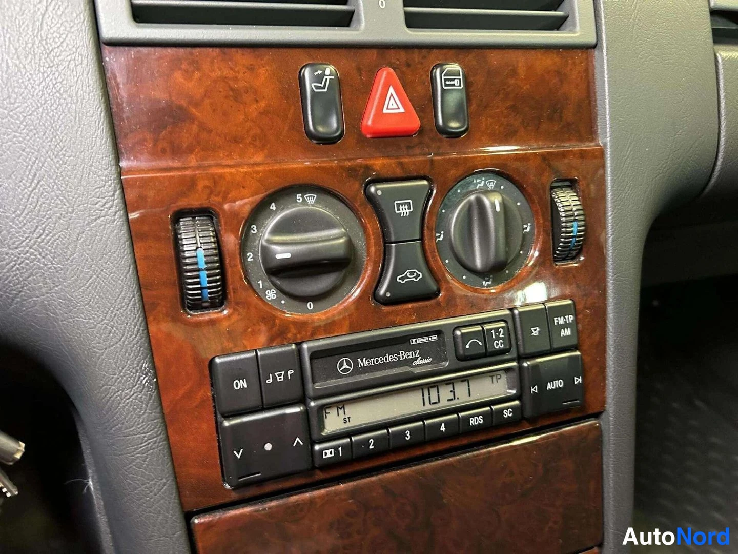 Edestä: Lähikuva Mercedes-Benz Classic -radiosta puidoitetussa keskikonsolissa: näyttö 103.7 FM ST, RDS- ja TP-merkkivalot, ON- ja AUTO-napit sekä ilmastoinnin säätimet