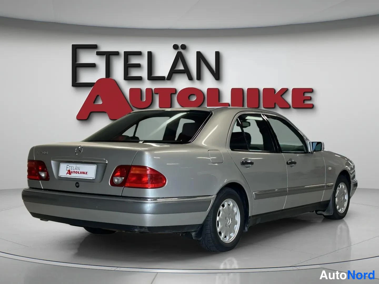 Takaviisto: Mercedes-Benz E-sarjan sedan takaa kolmannesnäkymässä, hopeanvärinen kori, Etelän Autoliike -taustakyltti.