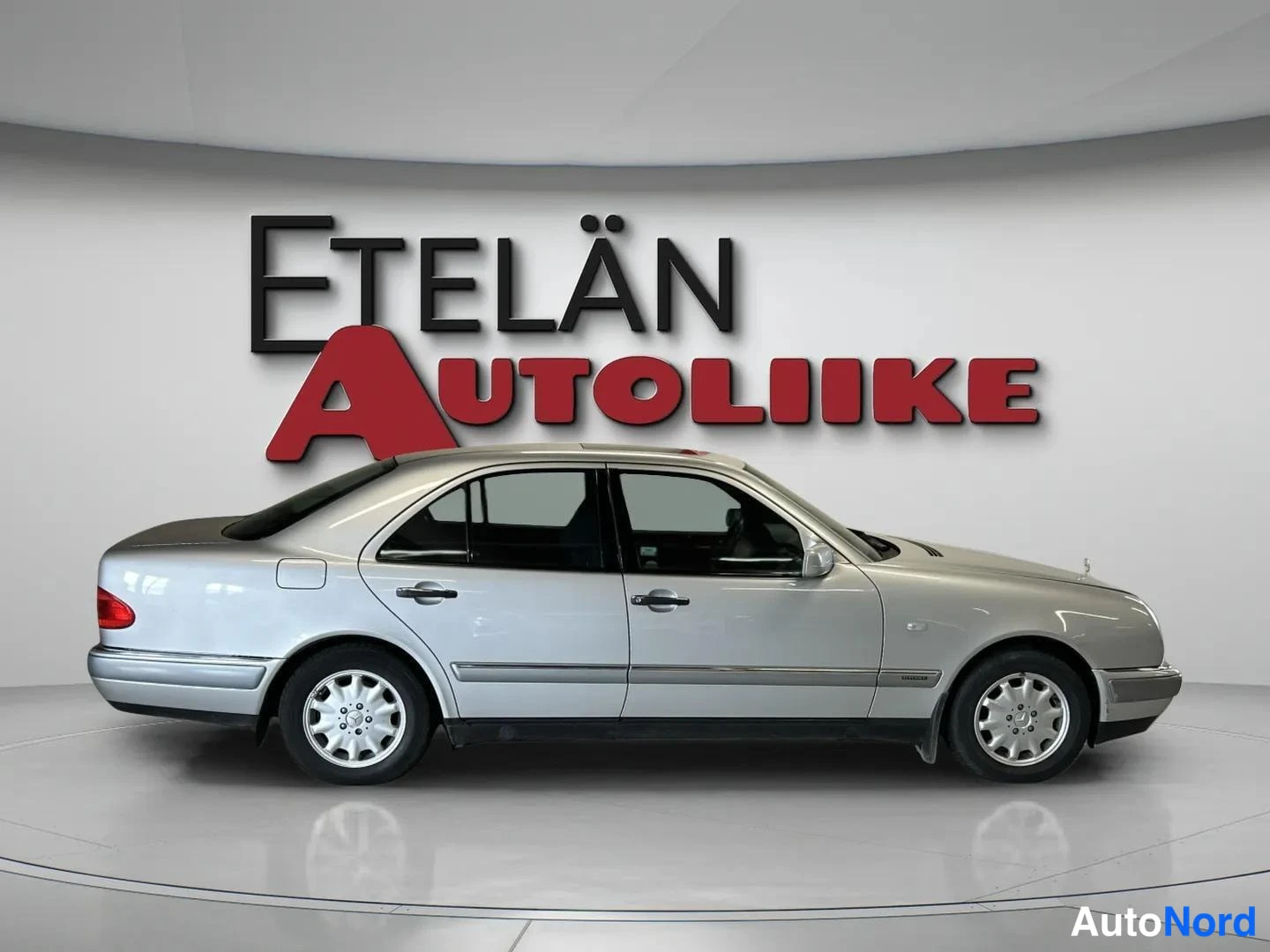Sivulta: Sivukuvassa hopeinen Mercedes-Benz E-sarjan sedan kuvattuna studiossa; taustalla Etelän Autoliike -teksti. Auto näkyy kokonaan kyljestään, takavalo osin näkyvis
