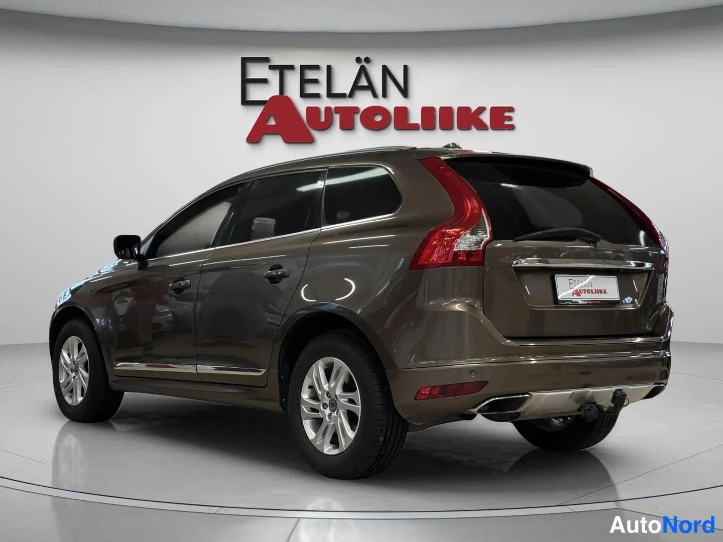 Takaviisto: Aud... — Takaosa ja vasen sivu näkyvä ruskeanharmaa Volvo XC60 -tyyppinen SUV studiovalaistuksessa, valkoinen tausta.