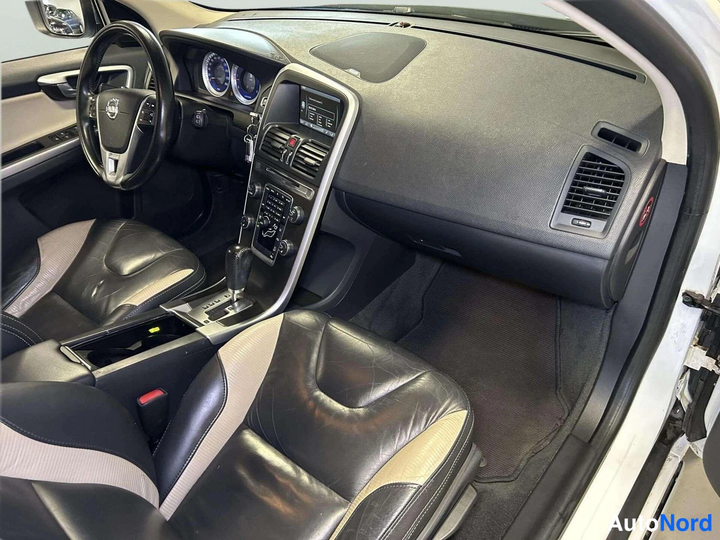 Etuviisto: Volvo XC60:n etummatila matkustajan puolelta kuvattuna: musta kojelauta, ilmastointilaitteet, automaattivaihteen valitsin ja musta-beiget nahkapenkit.