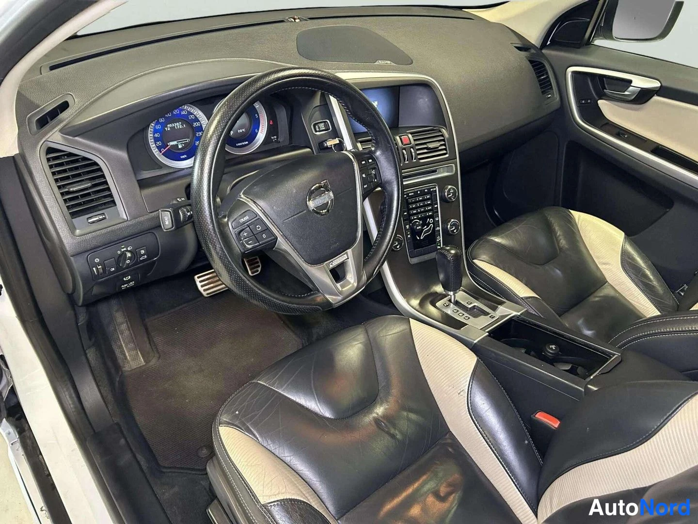 Etuviisto: Kuljettajan näkökulma Volvo XC60:n ohjaamoon: mittarit, Volvo-ohjauspyörä, automaattivaihteen valitsin ja musta-beiget nahkapenkit.