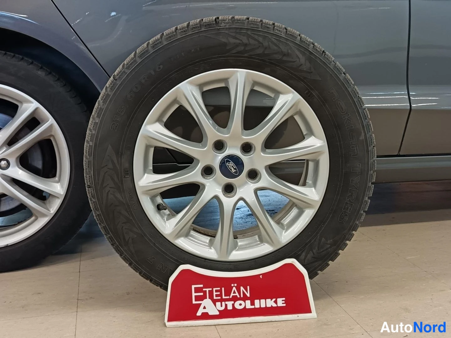Yksityiskohta: Alkuperäinen Ford-alumiinivanne ja Nokian talvirengas irrotettuna; renkaan sivussa lukee 215/60 R16 ja Nokian Tyres. Auto harmaa taustalla, Etelän Autoliike -ky