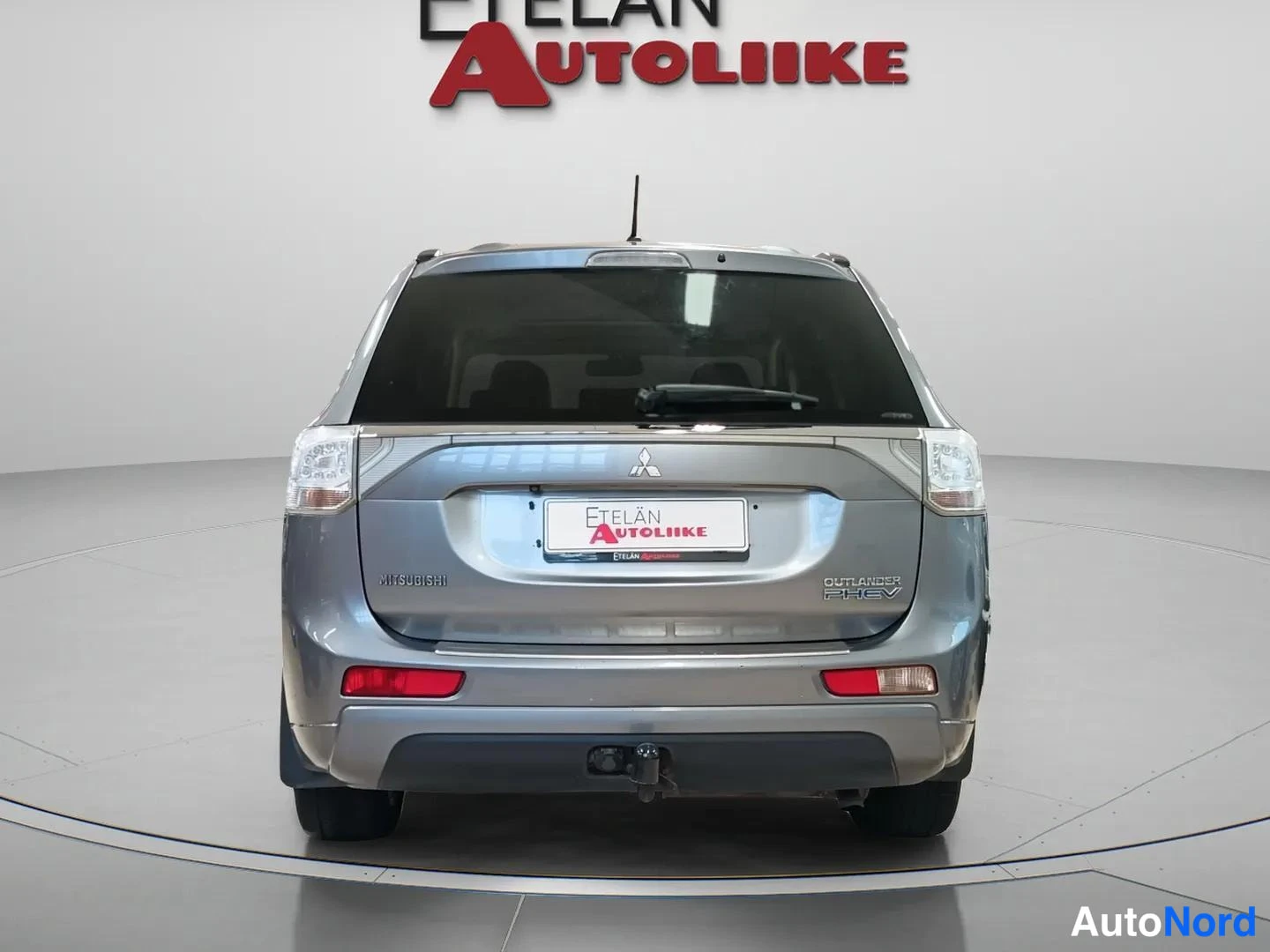 Takaa: Auton takanäkymä: harmaa Mitsubishi Outlander PHEV -takaluukki logoineen, takavaloineen ja hinauskoukulla varustettu puskuri.