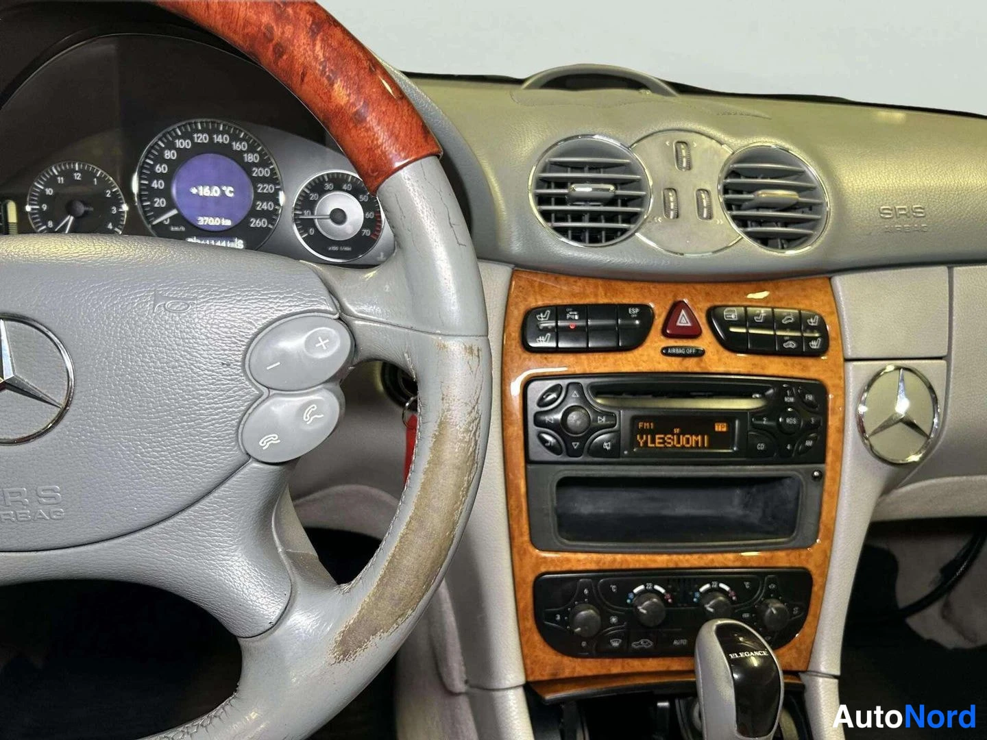 Muu: Mercedes-auton kojelauta ja keskikonsoli, puukuoripaneeleja ja radio näytöllä teksti YLESUOMI.