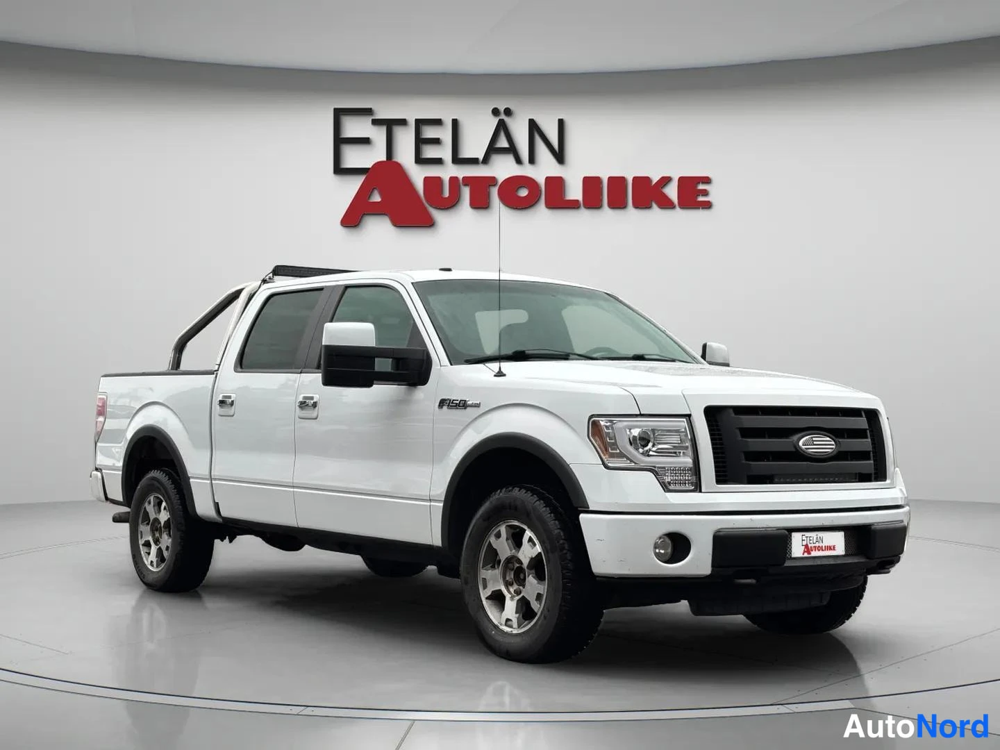 Etuviisto, nokka oikealle: Valkoinen Ford F-150 -lainen neliovinen avolava-auto kuvattuna kolmannenvuoronkulmasta, keula oikealla, studio-yleiskuvassa.