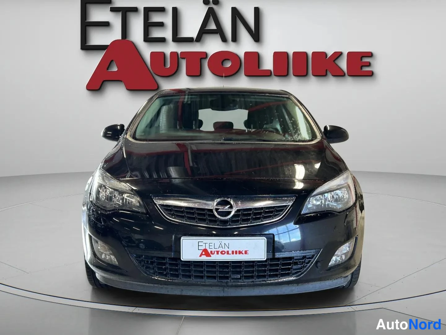 Edestä: Mustan auton suora edustanosto; etusäleikkö Opel-merkilla, ajovalot ja etupuskuri keskellä kuvaa.