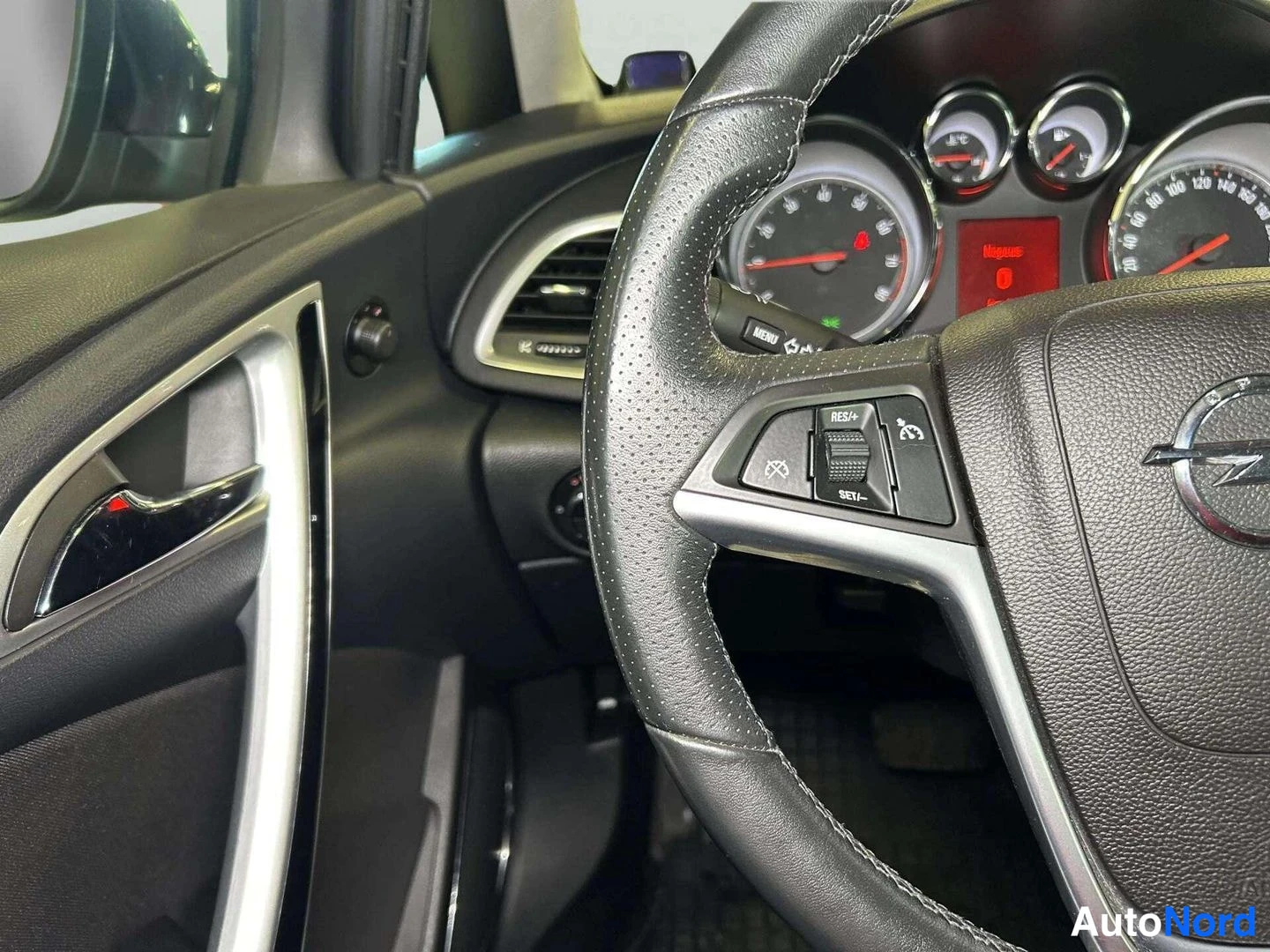 Sivulta: Rattipöydän lähikuva Opel-merkillä, nappiryhmät RES+ / SET– ja MENU-merkeillä, takaosassa mittarit.