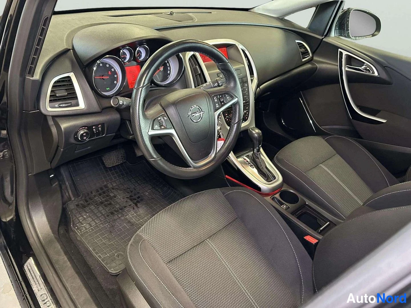 Edestä: Sisätilakuva: ohjaamo kuljettajan puolelta, Opel-ohjauspyörä, automatic-vaihteenvalitsin, kaksoset etuistuimet ja tummat matot.