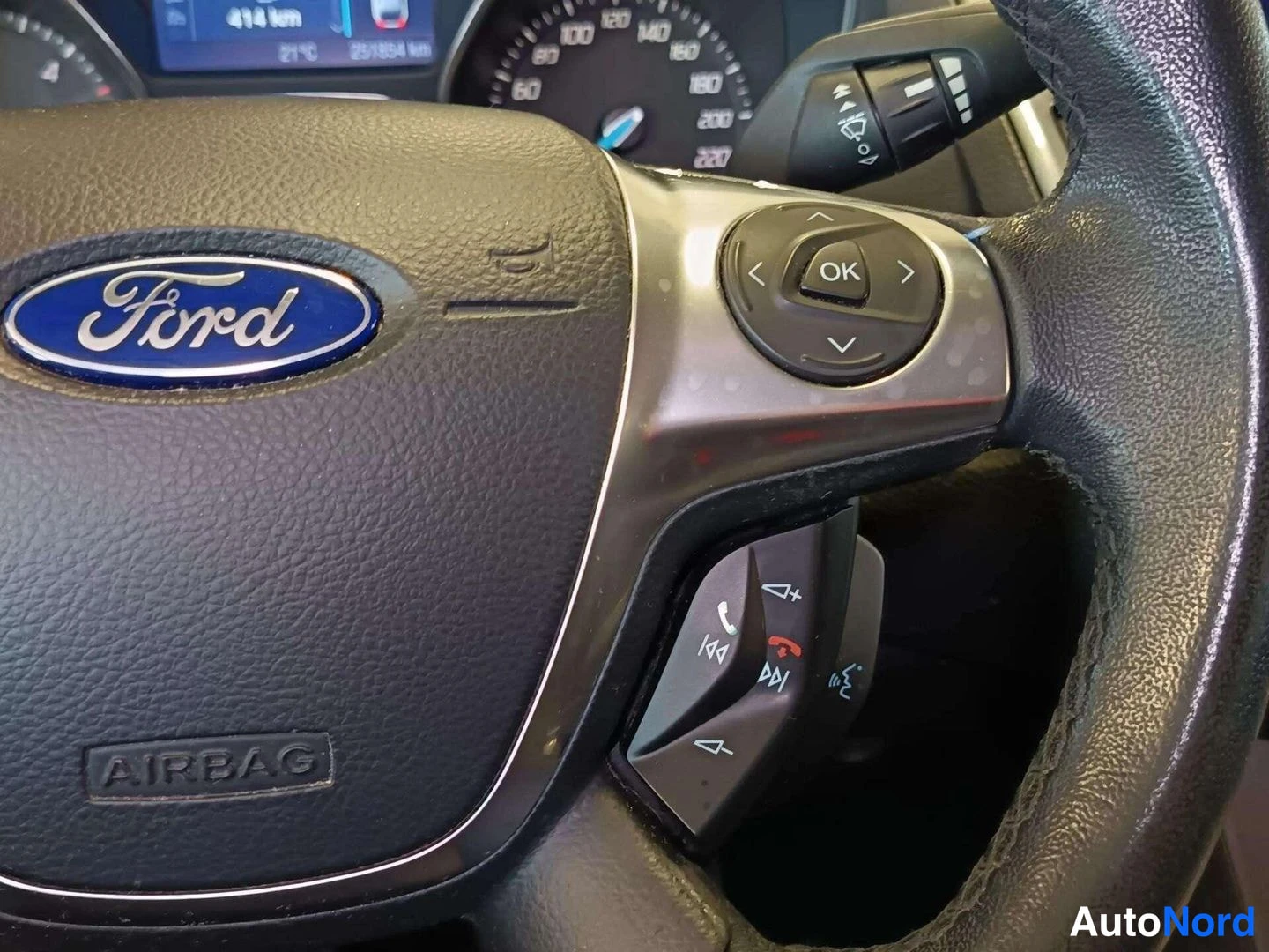 Muu: Lähikuva Ford-ohjauspyörästä: napassa AIRBAG-merkintä ja sininen Ford-logo, puhallimessa OK-painike sekä puhelin- ja ääniohjauspainikkeet. Taustalla pilkottaa k