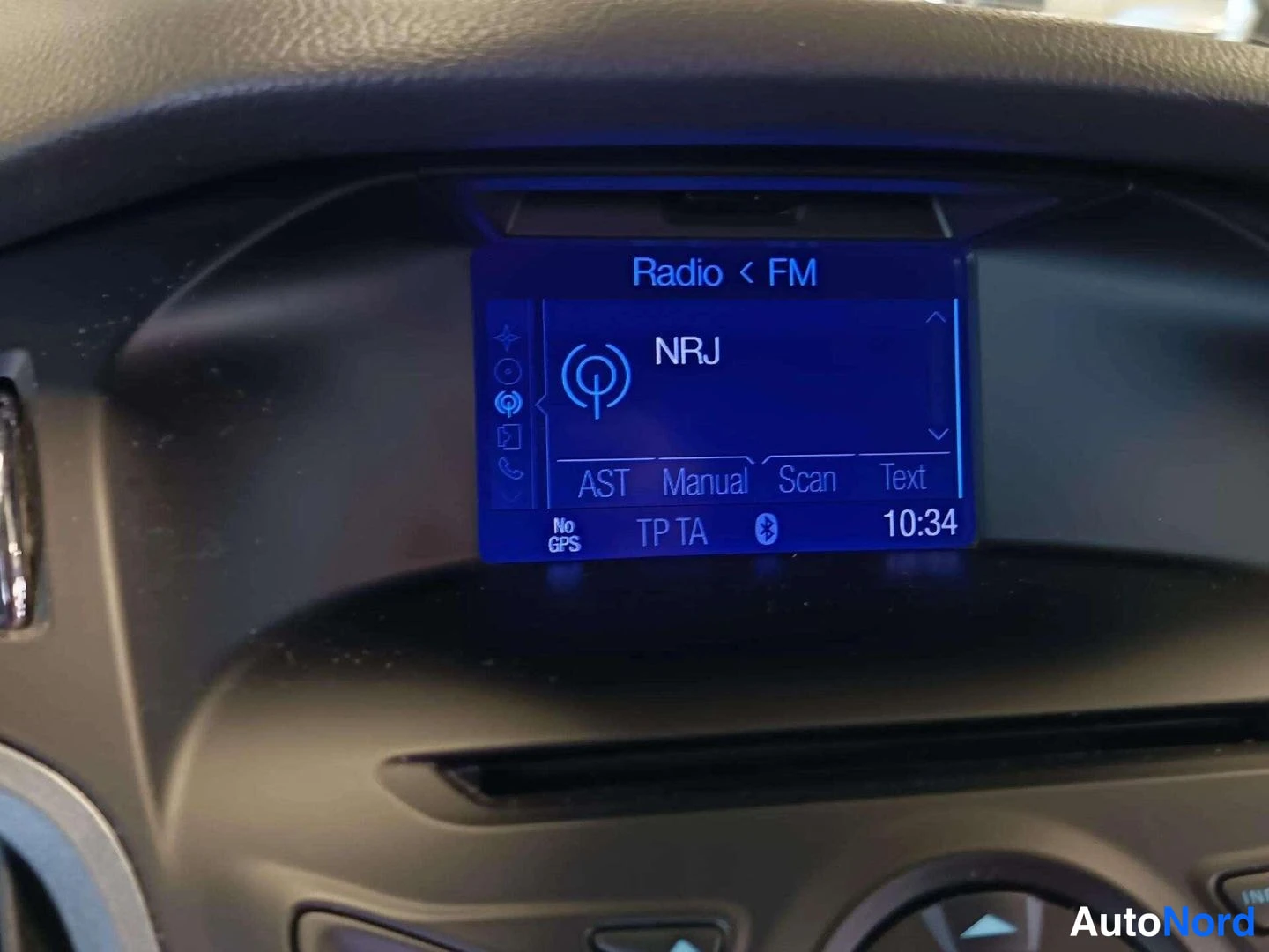 Muu: Auton kojelaudan radionäyttö, kanava NRJ, FM-radio, kello 10:34, Bluetooth-merkki näytössä.