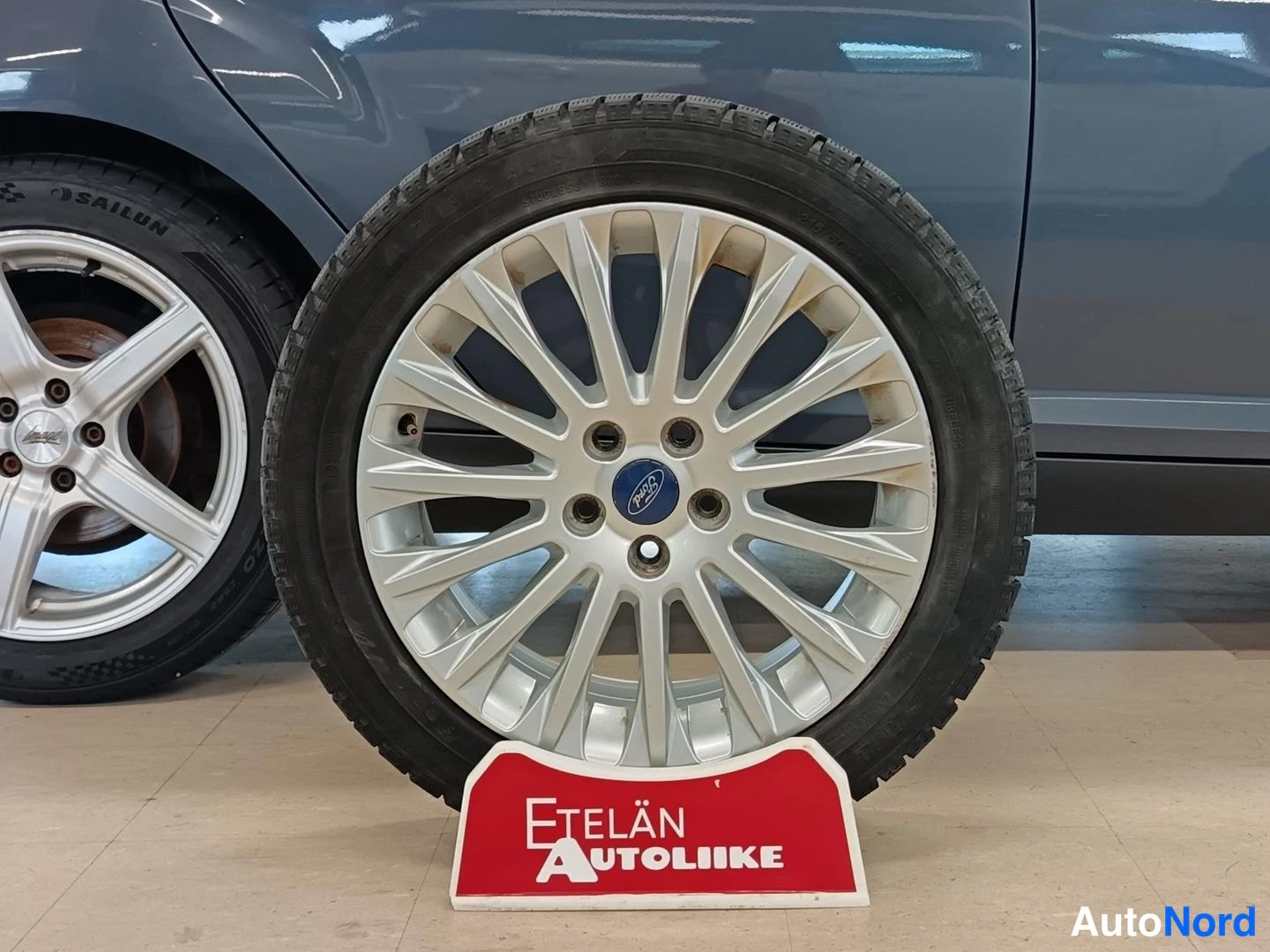 Yksityiskohta: Monisakarainen alumiinivanne Ford-logolla talvirenkaalla, edessä Etelän Autoliike -kyltti, taustalla auton sivu.