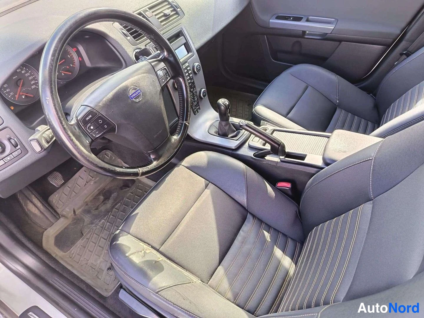 Muu: Volvo-merkkisen auton ohjaamo vasemmalta kuvattuna: ohjauspyörä, manuaalivaihtokeppi ja tummat etuistuimet.