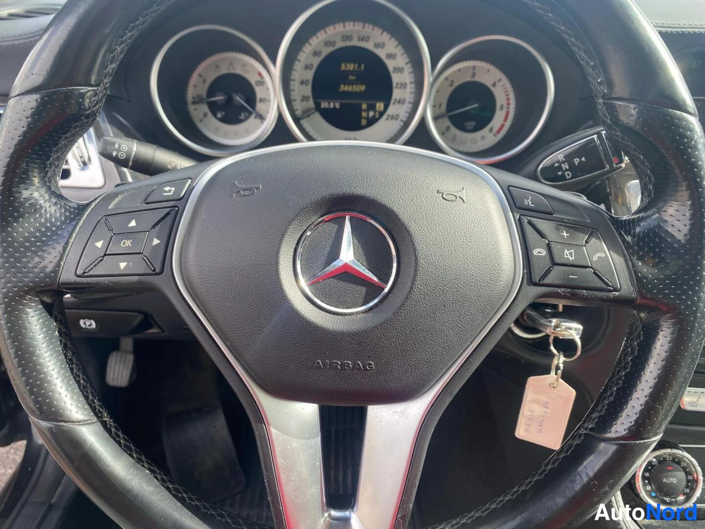 Sivulta: Auton sisätilat: Mercedes-merkkinen ohjauspyörä keskellä AIRBAG-teksti, takana kolme pyöreää mittaria ja digitaalinäyttö, jossa lukema 346509; avain roikkuu vir
