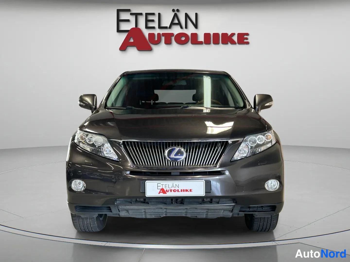 Edestä: Tummanharmaan Lexus RX -maasturin suora etunäkymä, grilli ja ajovalot studiotaustaa vasten.