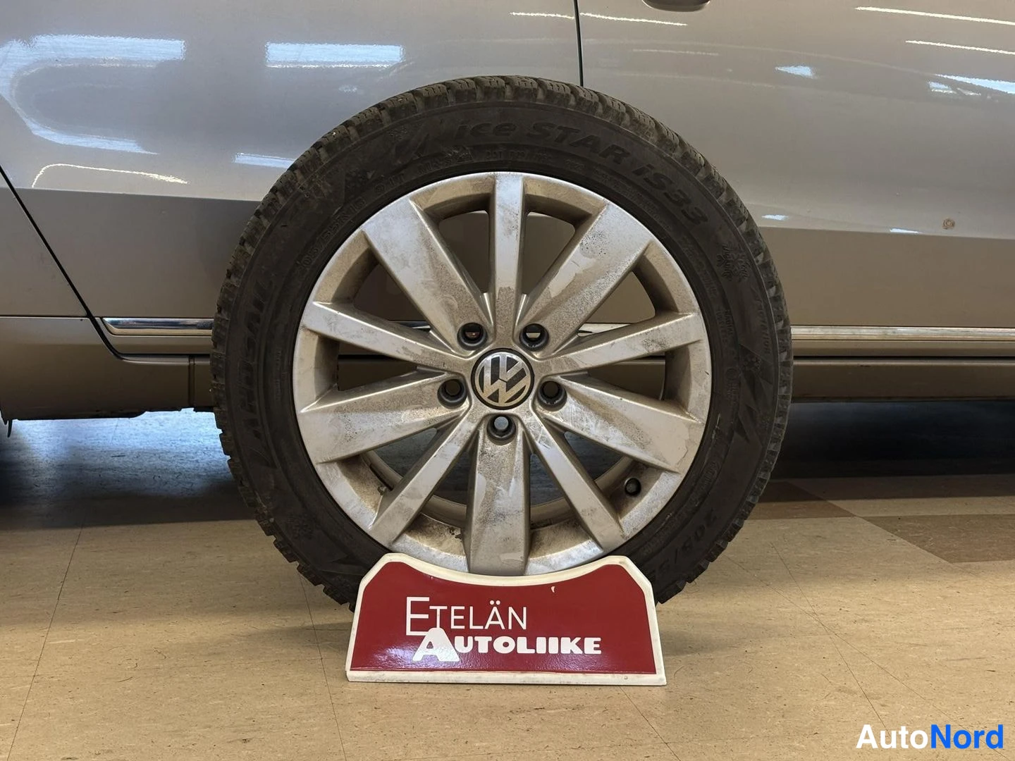 Muu: Volkswagen-merkkinen vanteellinen nokkarenkaallinen varapyörä seisoa lattialla punaisen Etelän Autoliike -kilpin päällä, auton kylki taustalla.