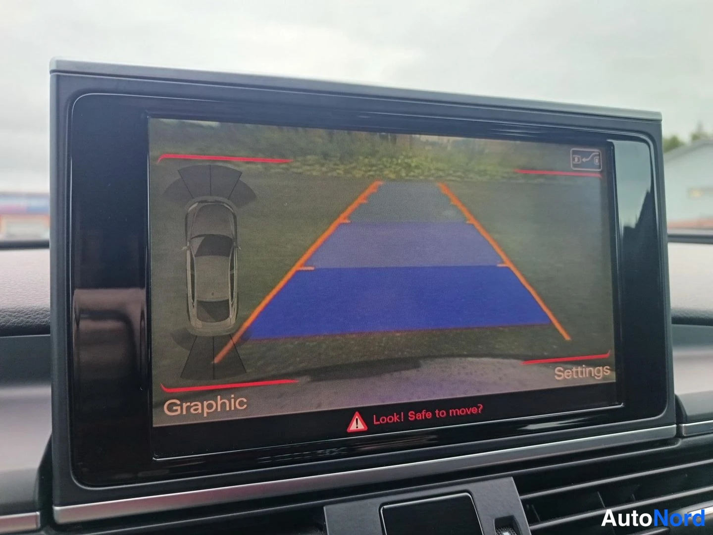Muu: Auton kojelaudan näyttö, jossa näkyy peruutuskameran kuva parkkiviivoilla ja tekstit Graphic, Settings ja Look! Safe to move?.