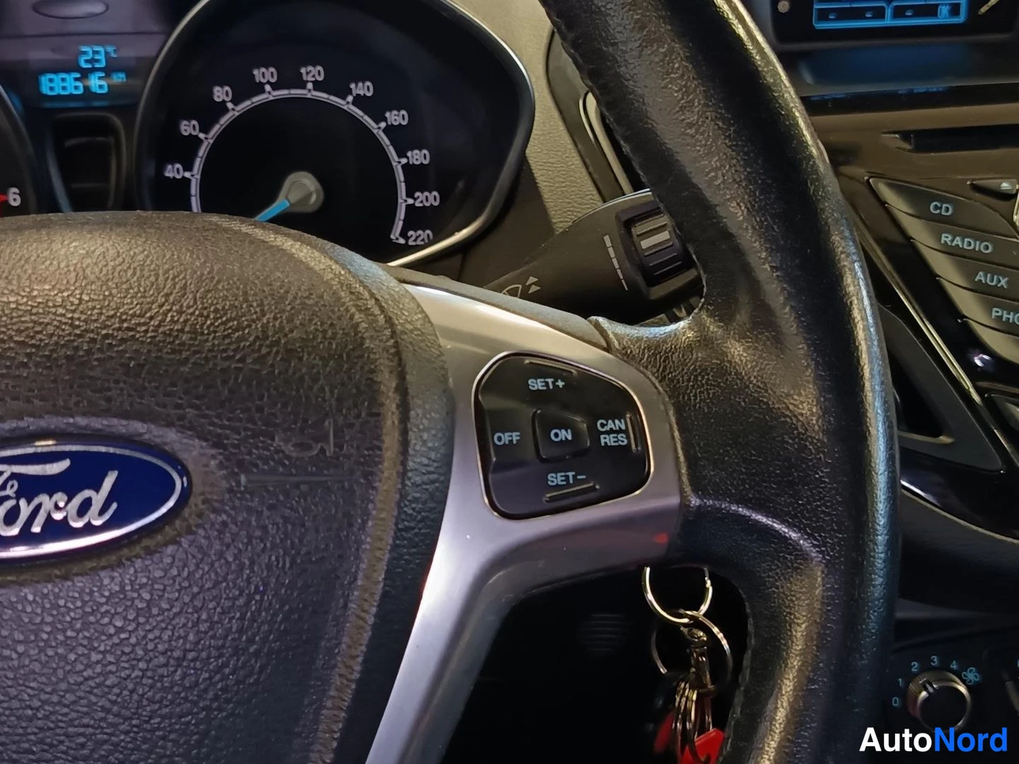 Muu: Ford-merkkisen auton ohjauspyörä lähikuvassa, nupissa Ford-logo ja vasemmalla puhelin- ja vakionopeudensäädinpainikkeet; takana osa mittaristosta.