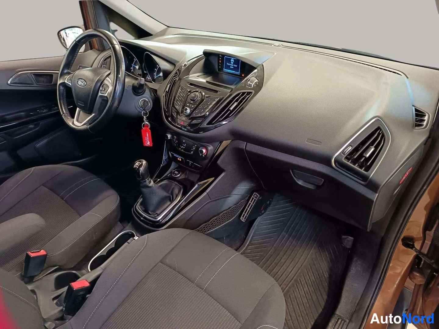 Ohjaamo: Front interior viewed from the open passenger side door. The black dashboard features a raised central display and air vents. A black gear stick is in the cente