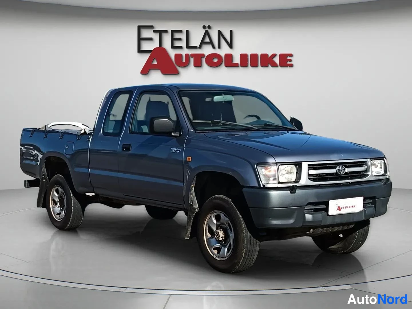 Etuviisto, nokka oikealle: Toyota Hilux-xtra-cab-pickup etuviistosta oikealta, tummansininen kori ja avolava peitolla.