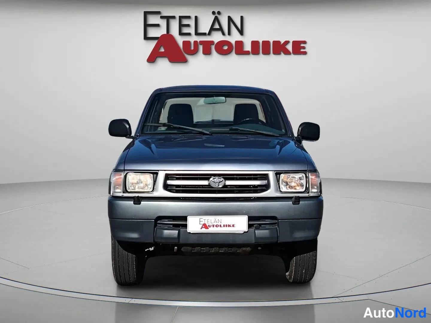 Edestä: Toyota Hilux suoraan edestä:Toyota-merkki tomaattorin, sumuvalot ja Etelän Autoliike-kyltti puskurissa.