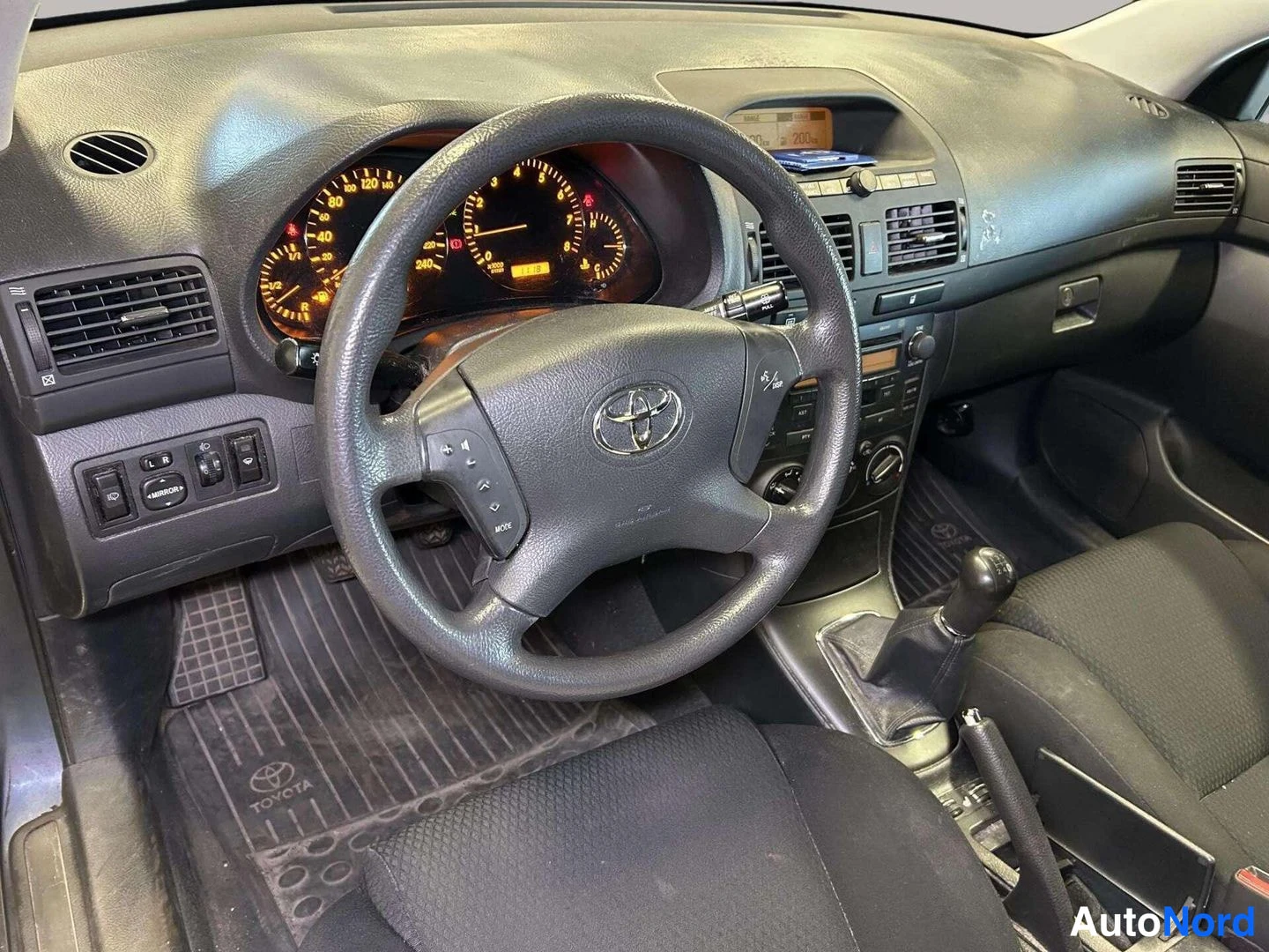 Muu: Toyotan ohjaamo vasemmalta kuvattuna: ratissa Toyota-embleemi, manuaalivaihdekeppi ja kojelaudan mittarit.