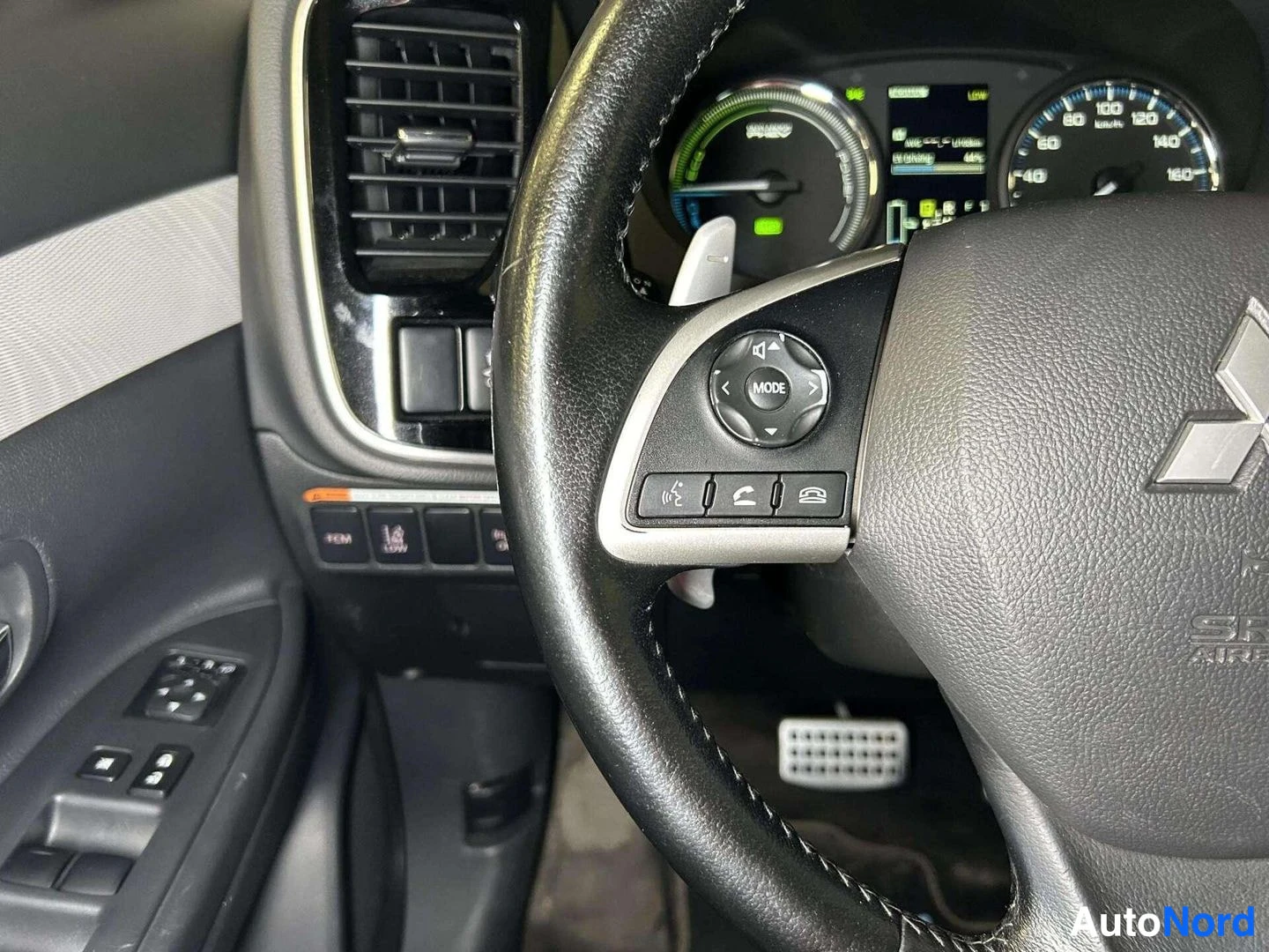 Sivulta: Ohjauspyörän ja kojetaulun lähikuva: Mitsubishi-merkki, MODE-nappi ja puhelinpainikkeet, hybridin mittaristo taustalla.
