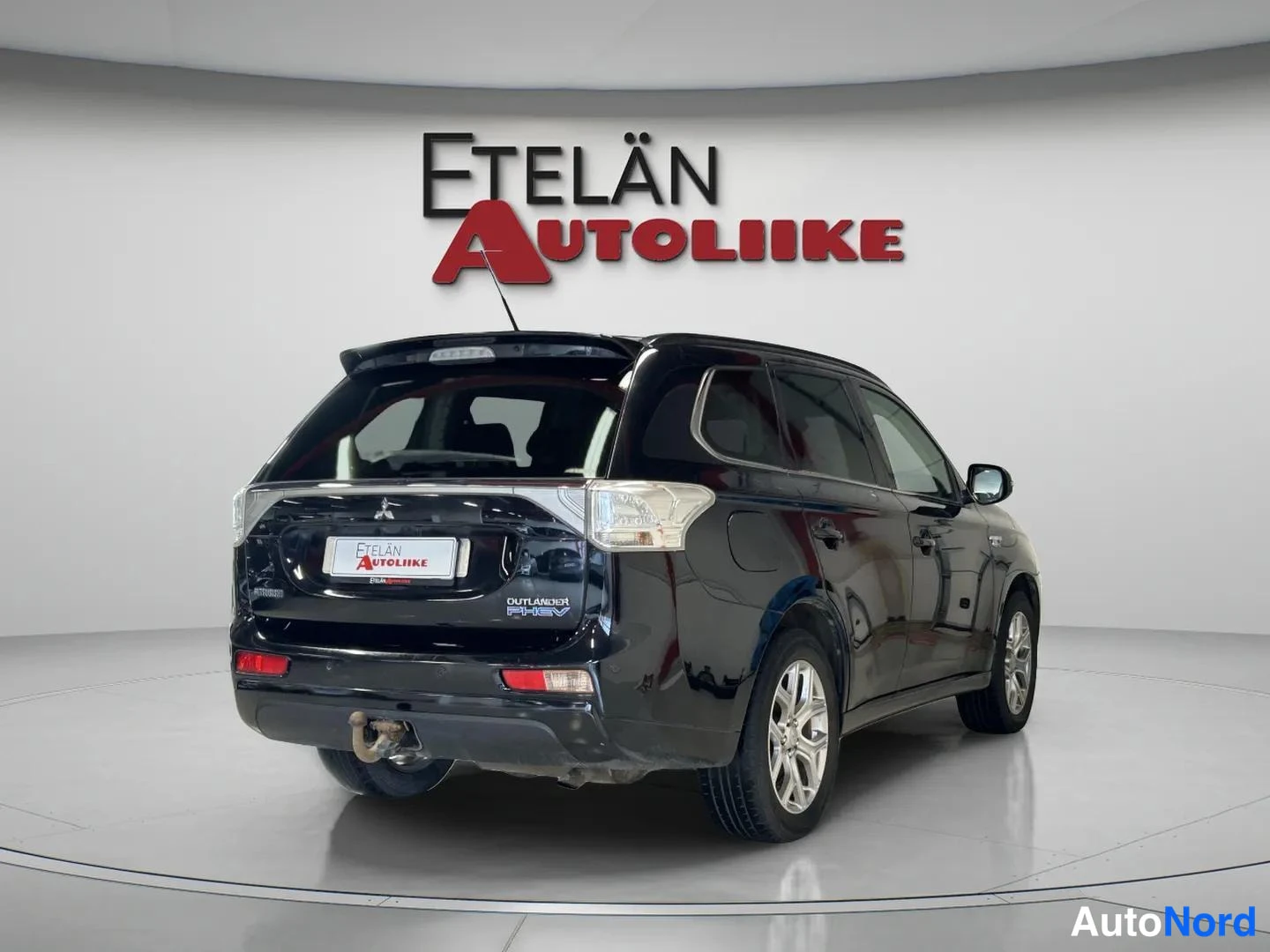 Takaviisto: Musta Mitsubishi Outlander kuvattu taka-vasemmalta kulmalta autoliikkeen tiloissa, takaluukissa OUTLANDER PHEV -teksti ja puskurissa vetokoukku.