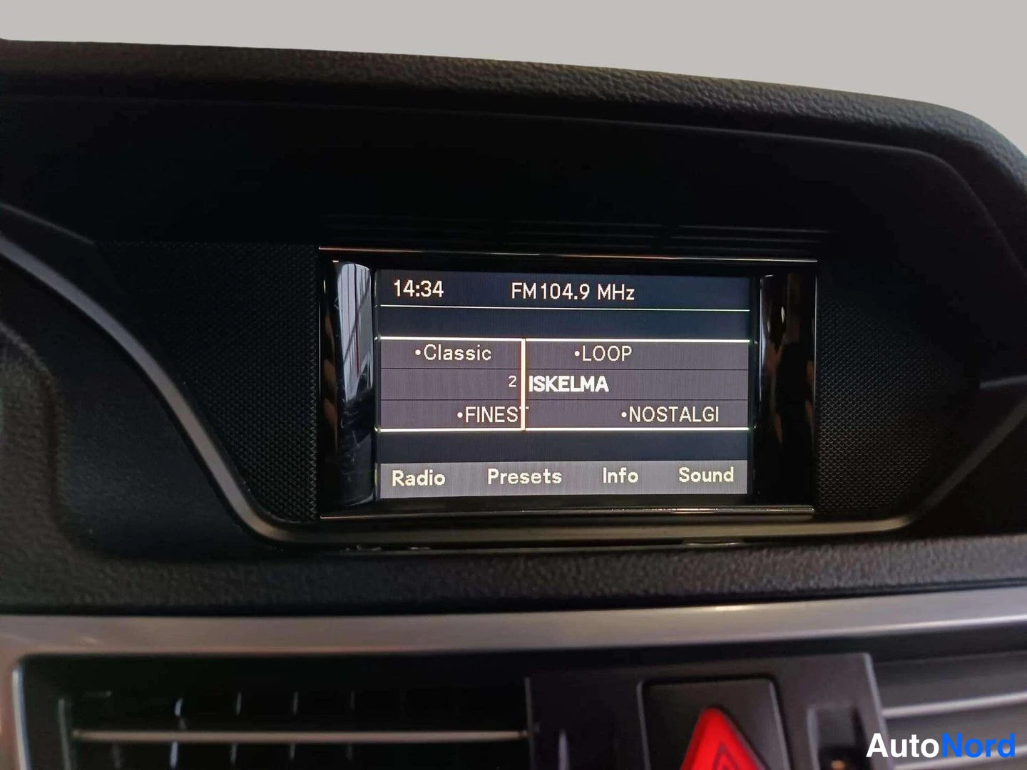 Yksityiskohta: Auton kojelaudan näyttö näyttää radio FM104.9 MHz -kanavan ja eri vaihtoehdot. Näytön ympärillä on tummaa muovia.