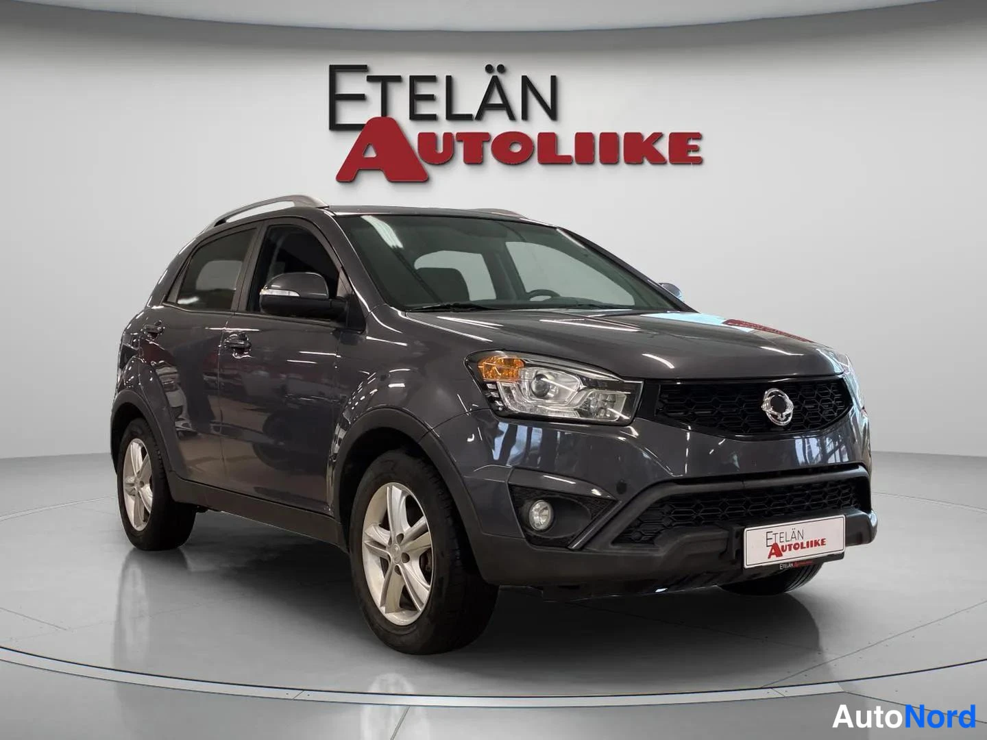 Etuviisto, nokka oikealle: Harmaa korkeakoriin henkilöauto studiotyttö­puistossa, etukulmasta oikealta kuvattuna, Etelän Autoliike -logo taustalla.
