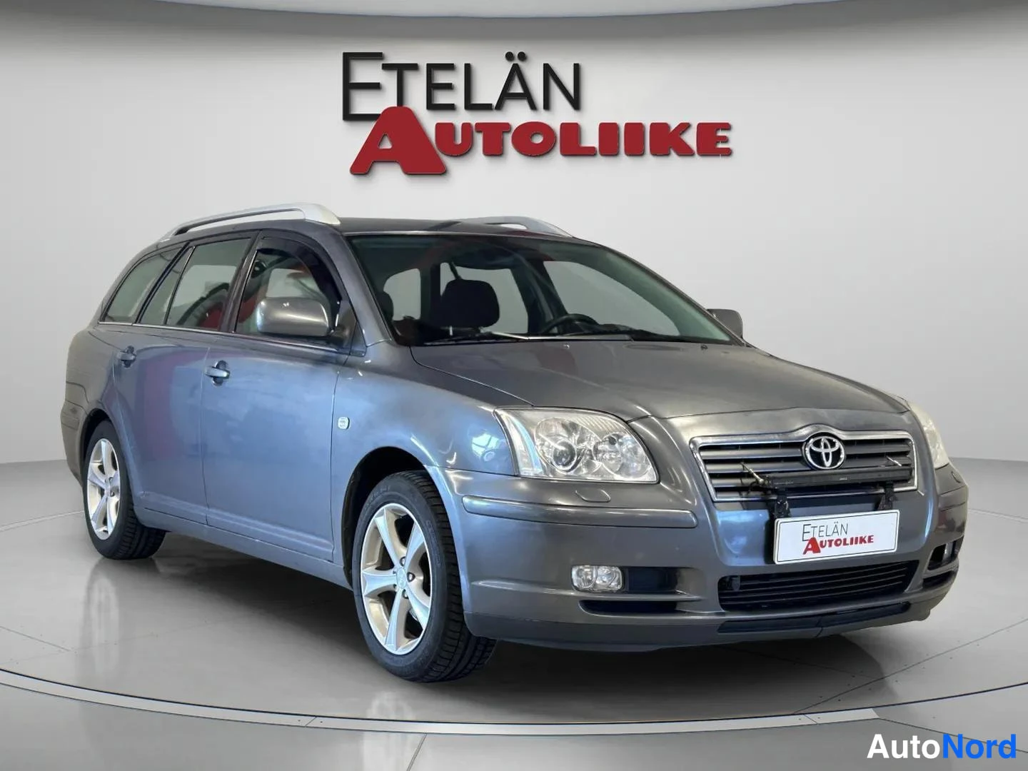 Etuviisto, nokka oikealle: Autoliikkeen esittelytilassa harmaa Toyota Avensis -farmariauto kuvattu kolmasosa-etesuvkulasta, keulan osoittessa oikealle.