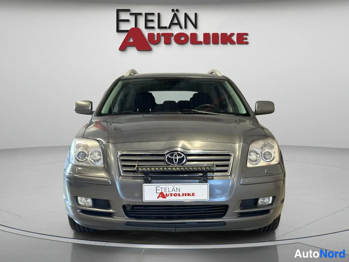 Edestä: Suora etunakyma harmaasta Toyota Avensis -farmariautosta liikkeen esittelytilassa, etupuskuri ja ajovalot nakytavissa.