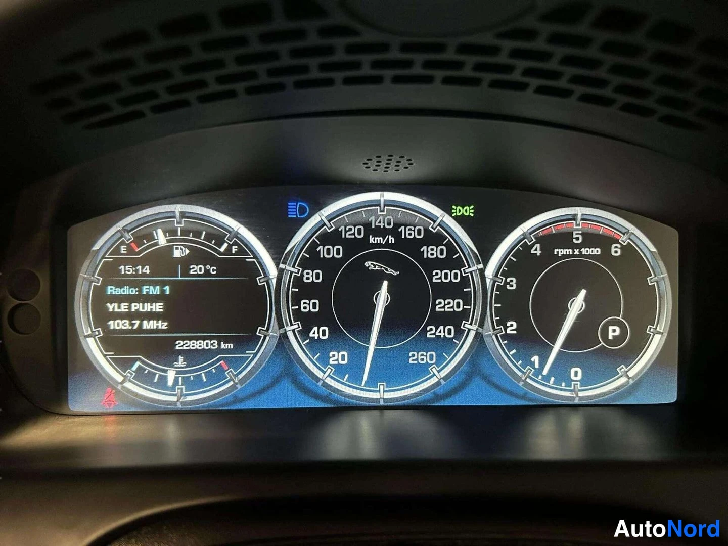 Yksityiskohta: Mittariston näyttö: nopeusmittari 260 km/h asti, kierroslukumittari P-vaihteella, polttoainemittari ja radiotiedot 'YLE PUHE 103.7 MHz', matkamittari 228803 km.