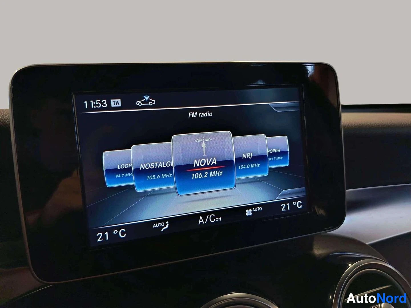 Muu: Auton keskinäyttö FM-radio-näkymässä, valittuna asemana NOVA 106.2 MHz; näytön alareunassa ilmastoinnin A/C ON ja AUTO sekä lämpötila 21 °C.