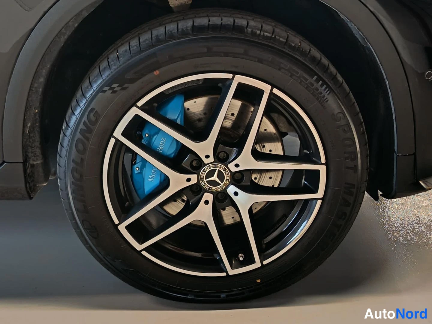 Yksityiskohta: Mustan Mercedes-henkilöauton takapyörän lähikuva: viisipoikkeinen kevytmetallivanne, sininen Mercedes-Benz-jarrusatula ja suurikokoinen jarrulevy näkyvät renkaa