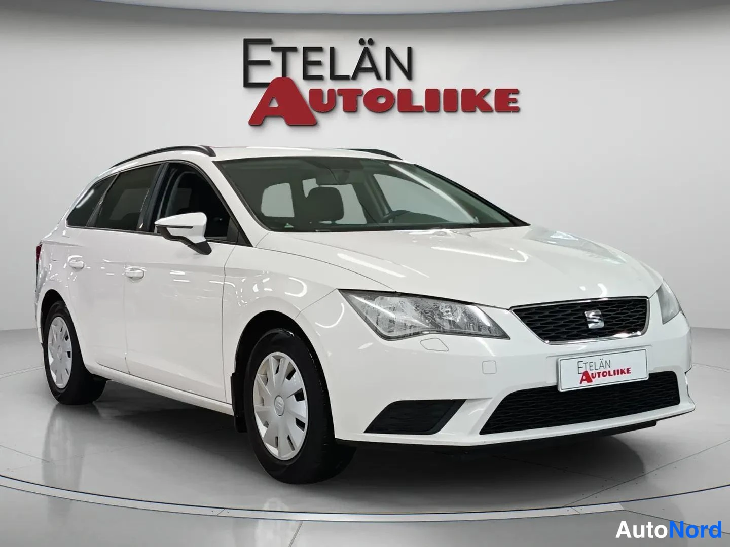 Etuviisto, nokka oikealle: White estate car photographed from a front three-quarter angle, nose pointing to the right, in a dealership studio with dealer logo on the wall.