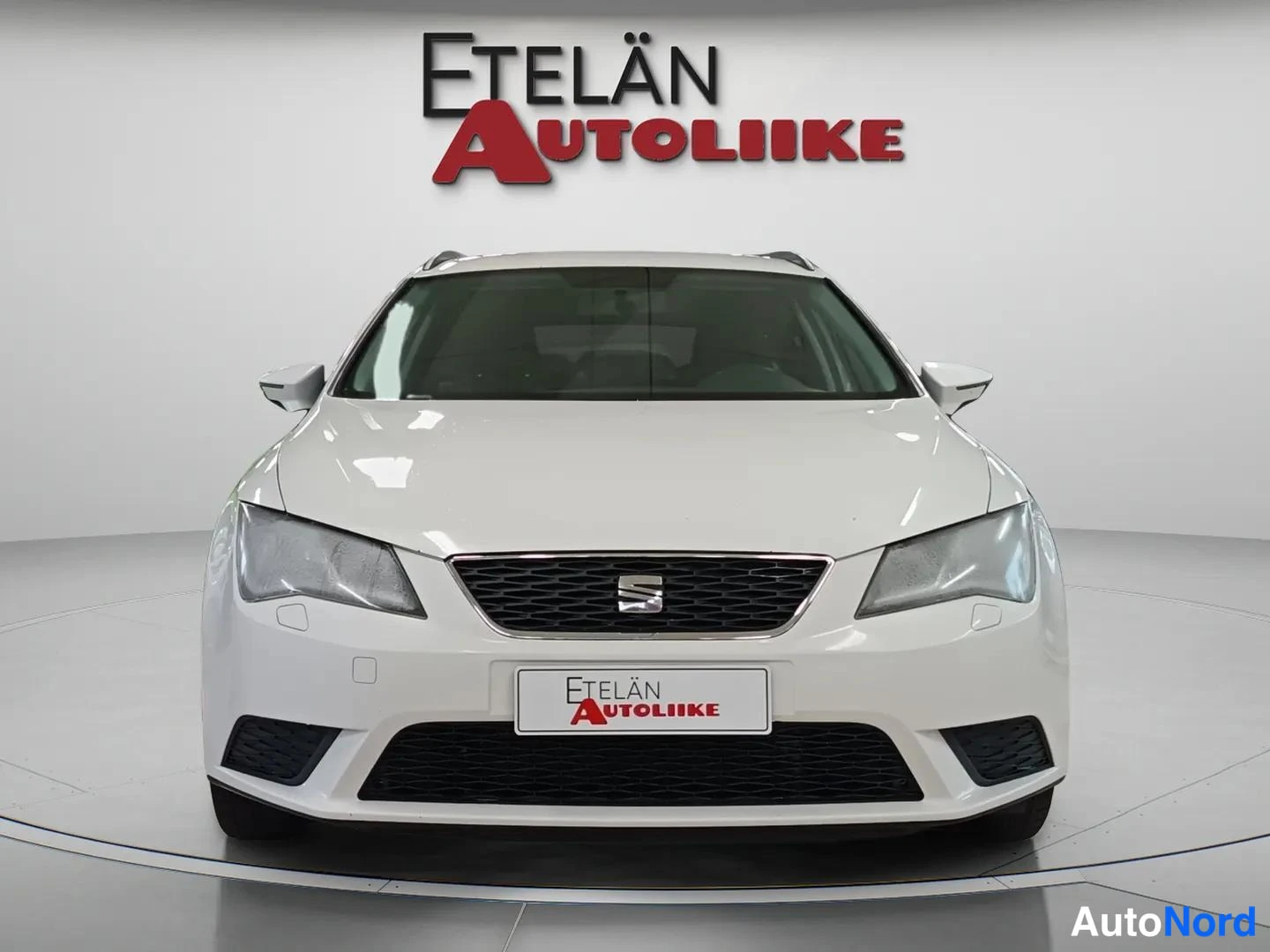 Edestä: Front view of a white estate car showing grille, headlights and bumper in a dealership studio.