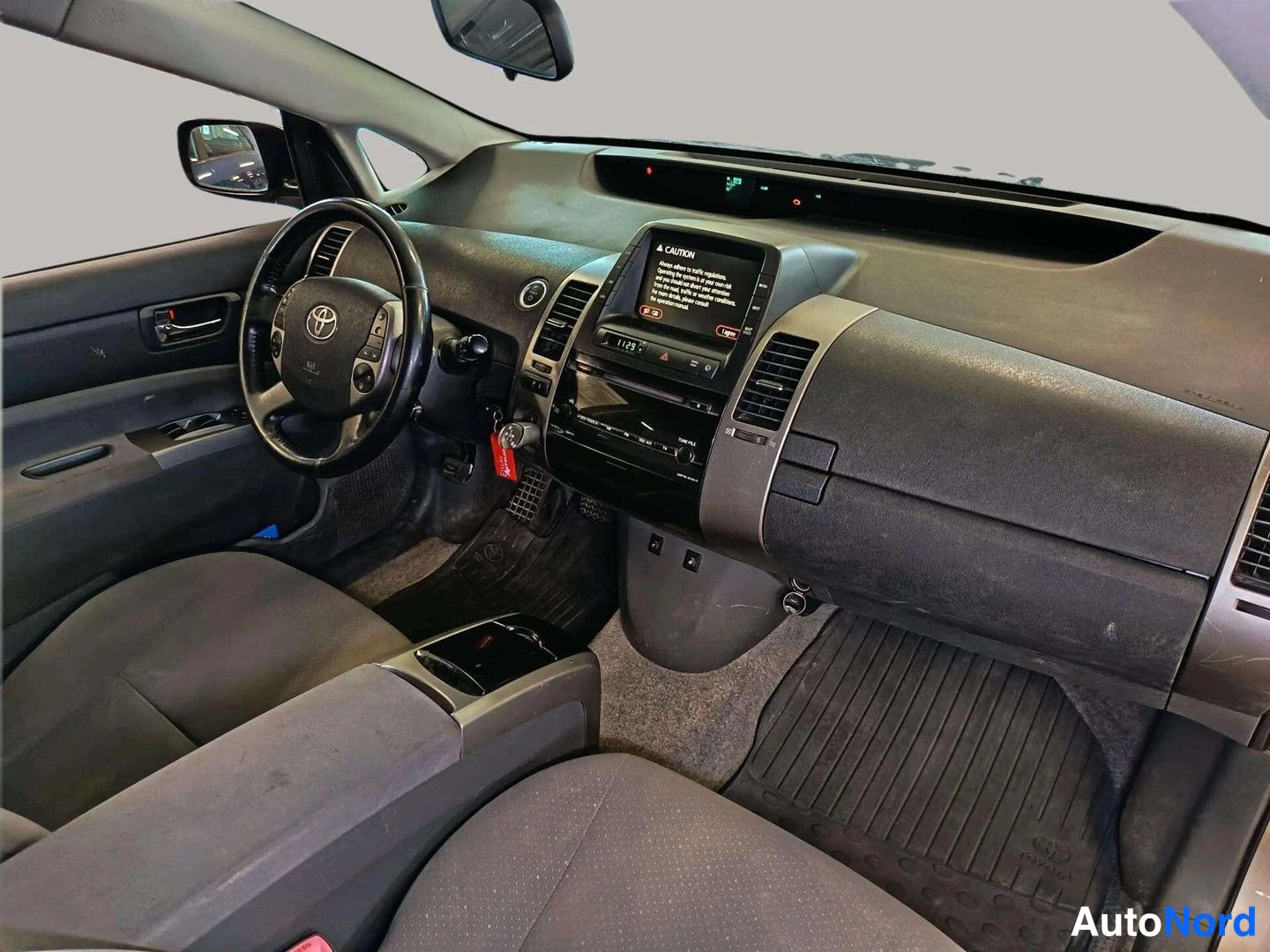 Muu: Toyota Priuksen ohjaamo matkustajan puolelta kuvattuna: ohjauspyörä, keskikonsolin infotainment-näyttö,Automatic-ruisku ja tummat matot.