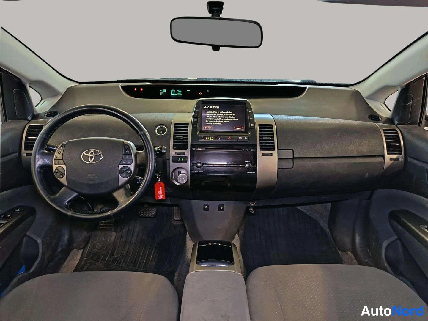 Muu: Toyota Priuksen kojelauta suoraan edestä kuvattuna: Toyota-ohjauspyörä, hybridi-ilmaisinryhmä tuulilassin alla, keskikonsolin CAUTION-näyttö ja punainen avainlä