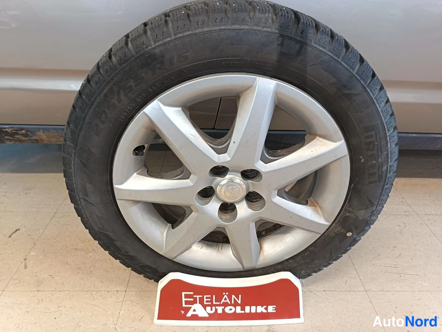 Yksityiskohta: Vaihtopyyrä lattialla: hopeinen alumiinivanne ja Pirelli-talvirengas (205/55 R16), taustalla punainen Etelän Autoliike -kyltti.
