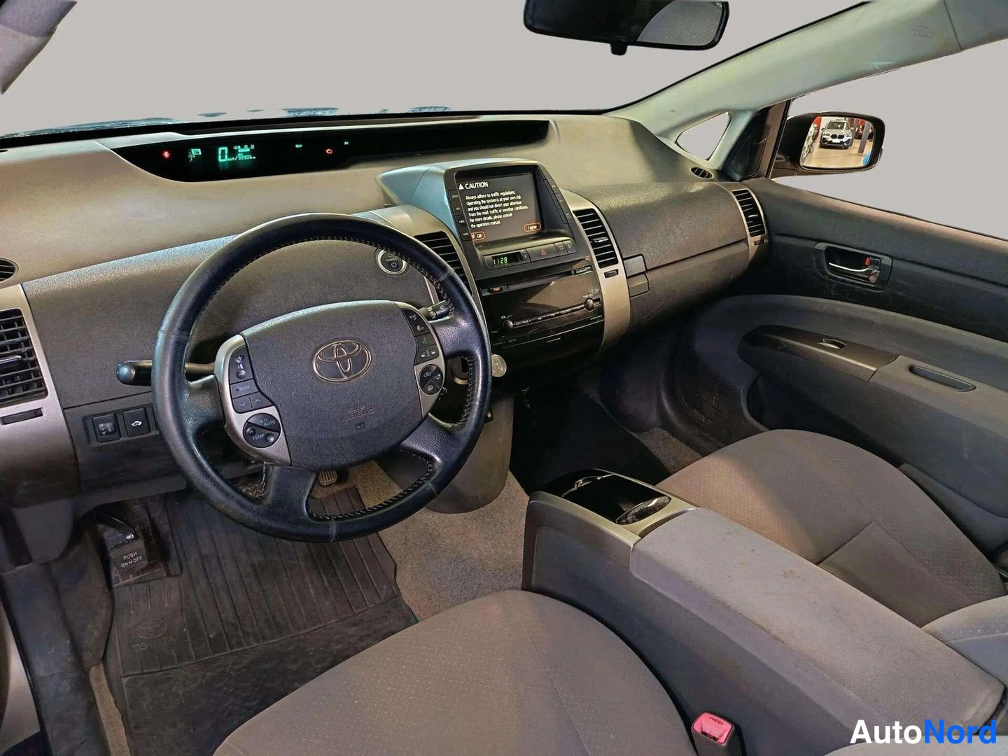 Muu: Toyota Priuksen ohjaamo etuistuimelta kuvattuna: hallintapaneeli, Toyota-ohjauspyörä, keskikonsolin näyttö ja tummat kangasistuimet.