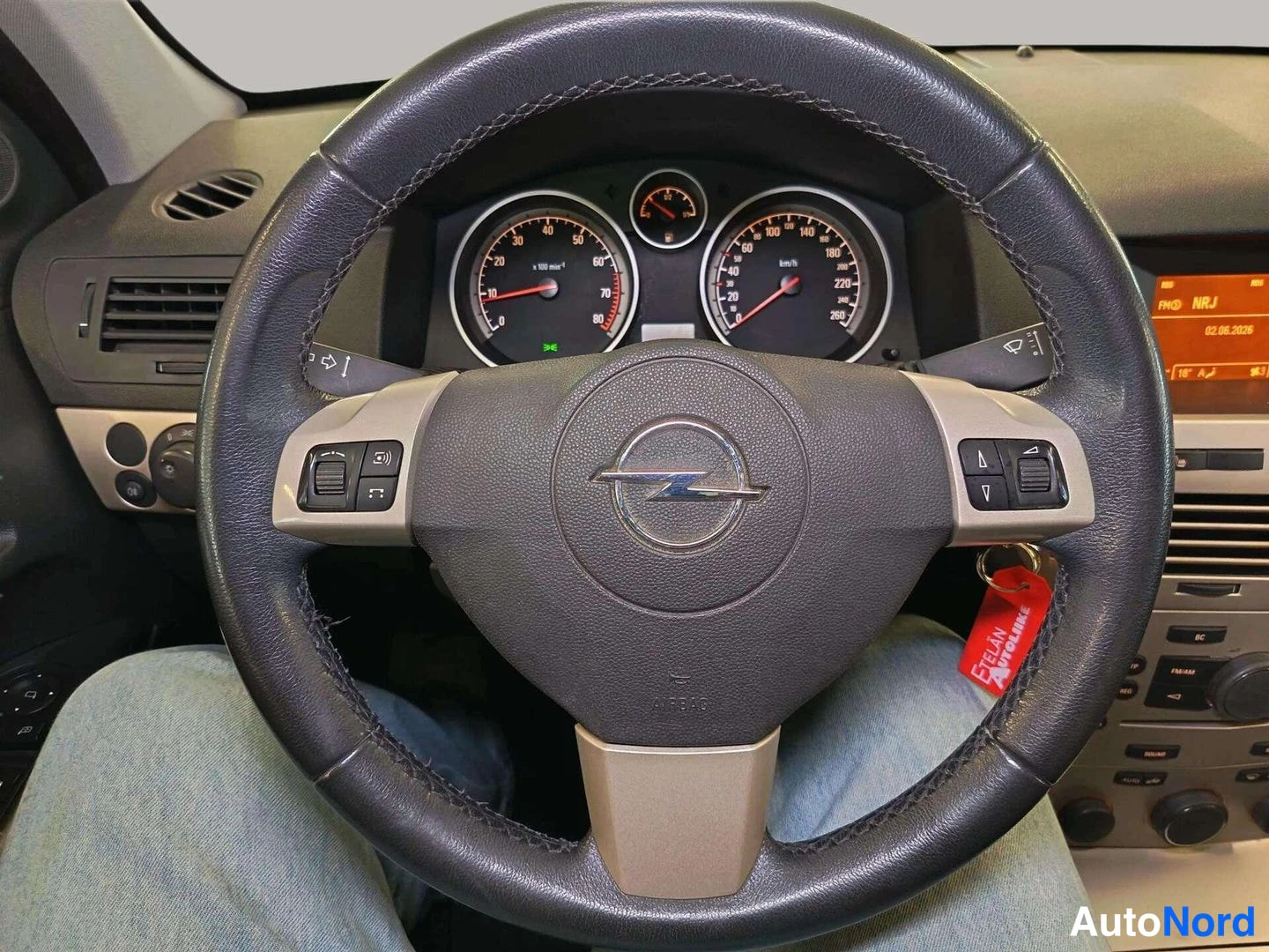 Yksityiskohta: Opel-ohjauspyörä lähikuvassa, puhelin- ja nuolipainikkeet, mittaristo näkyy taustalla; punainen avainnenkkuli roikkuu sytytyslukossa.