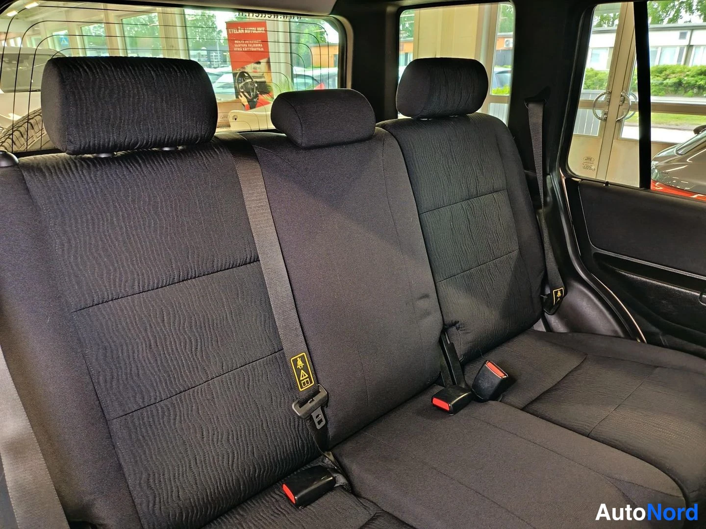 Takaistuimet: Rear bench seat with three-position headrests and dark fabric upholstery; ISOFIX anchors visible on the outer seat positions indicated by yellow warning labels;