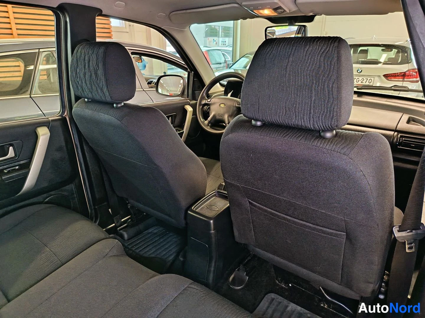 Ohjaamo: Interior view from rear looking forward to front seats showing black textured fabric seats; steering wheel and side mirror visible through dashboard gap; light