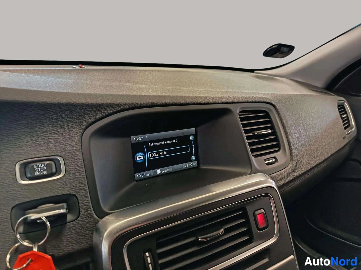 Yksityiskohta: Kojelaudan keskusta: START STOP ENGINE -painike, radio- ja puhelinvalikko näyttävä infotainment-näyttö, ilmastointisuuttimet ja avaimet tepassä.
