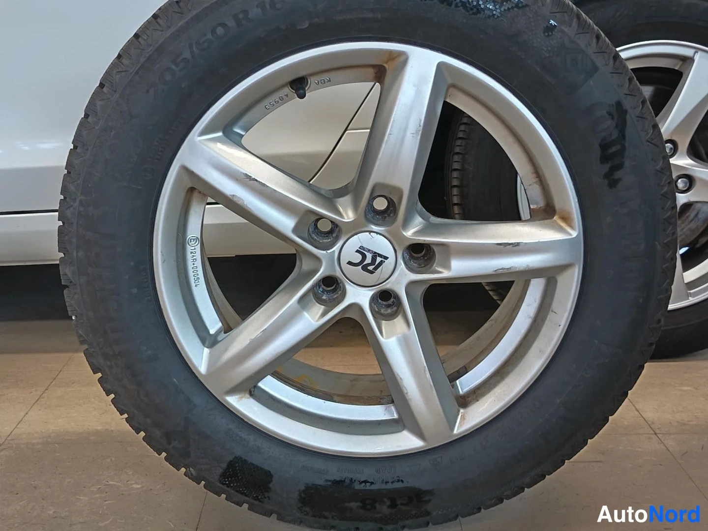 Sivulta: Irrotettu talvirengas 205/60 R16 hopeisessa viisikuvioisessa RT-vanteessa seisoo lattialla auton vieressä.