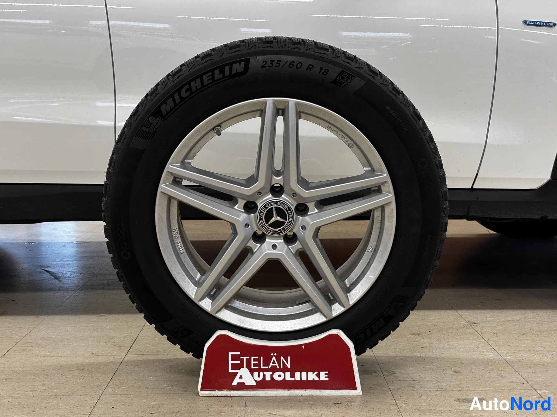 Sivulta: Vaalean autocen kylki ja pyörä lähikuvassa: hopeinen viisipuolainen Mercedes-vanne, Michelin-talvirenkaan kyljessä lukee MICHELIN 235/60 R 18. Etualalla punaine
