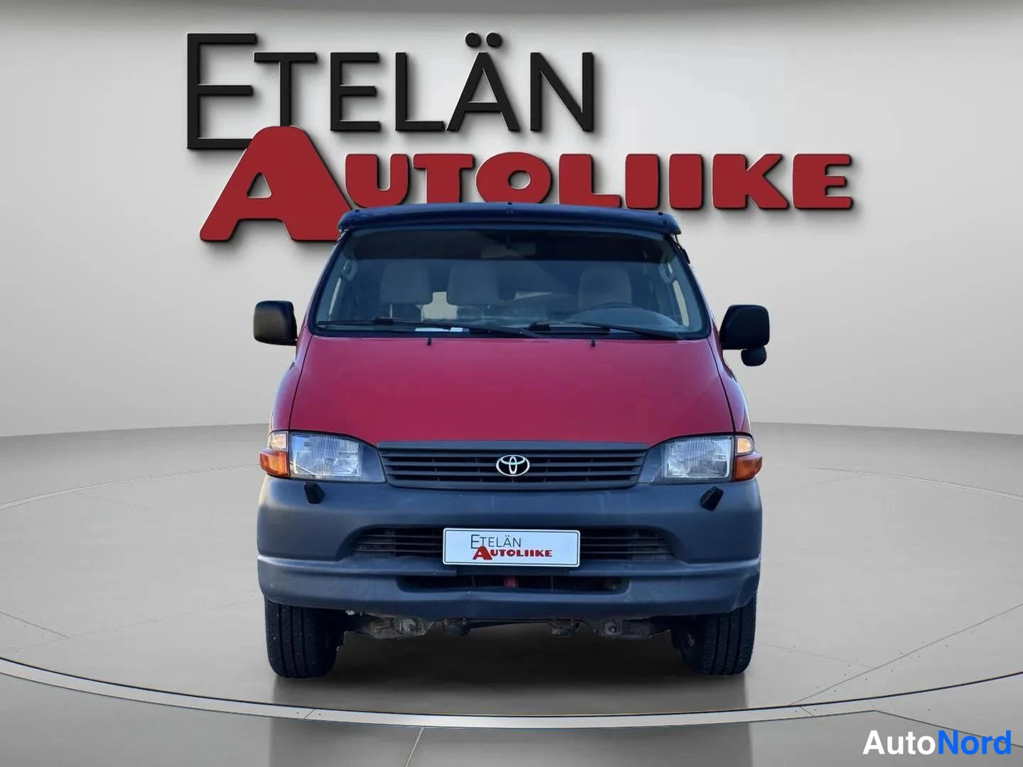 Edestä: Punaisen pakettiauton etänäkymä, Toyota-merkki etusäleikössä studiotaustaa vasten.