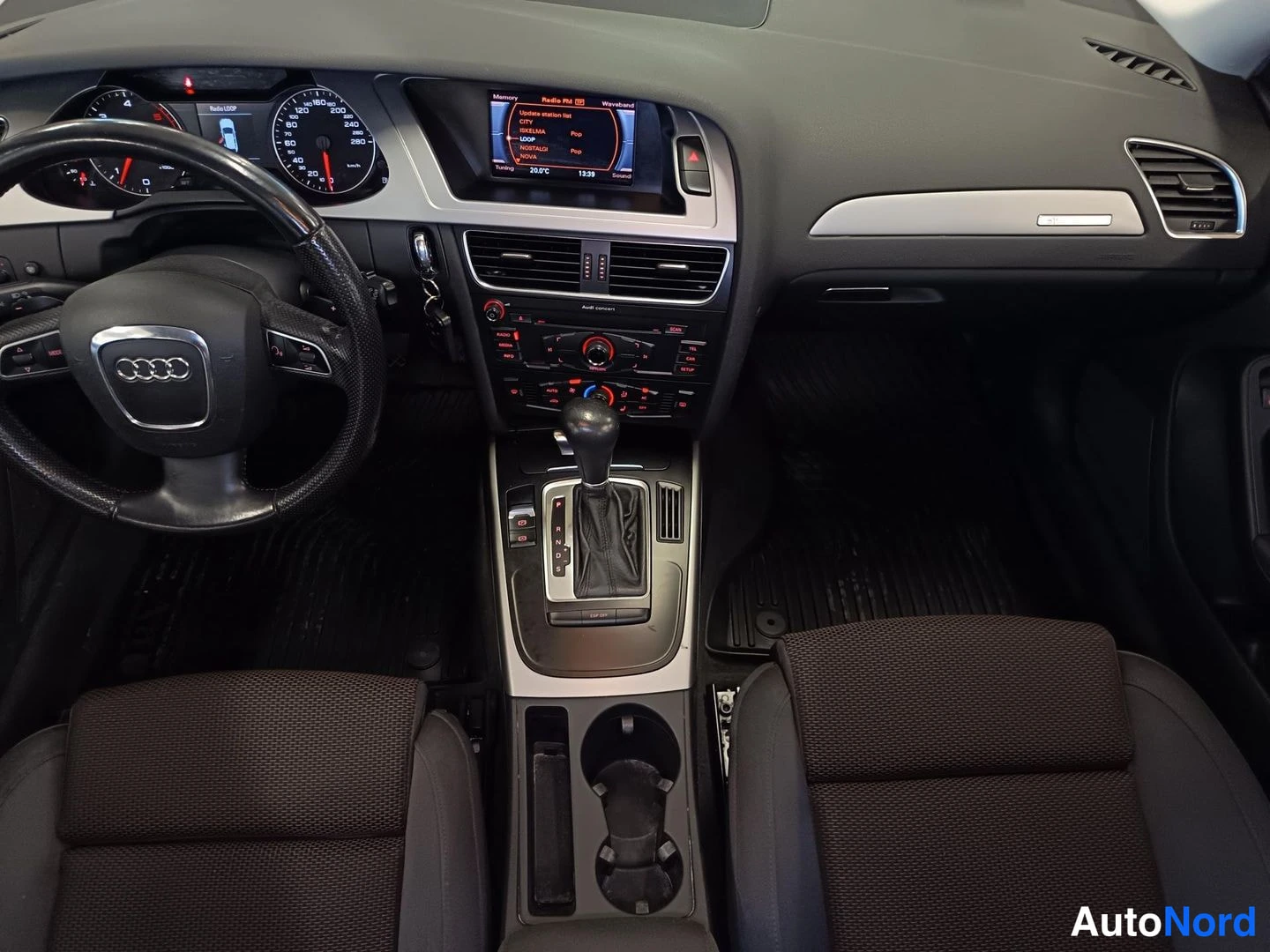 Edestä: Yläviistokuva kojelaudasta: Audi-ohjauspyörä, mittaristo, radion valikko näyttävä infotainment-näyttö, automaattivaihteen valitsin ja kaksi etuistuinta.