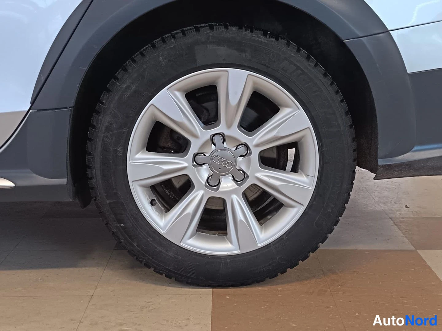 Muu: Audi A4:n pyörä lähikuvassa: hopea monipuolinen Audi-vanne mustalla kitkatalvirenkaalla, auton kylki ja musta lokasuojus taustalla.